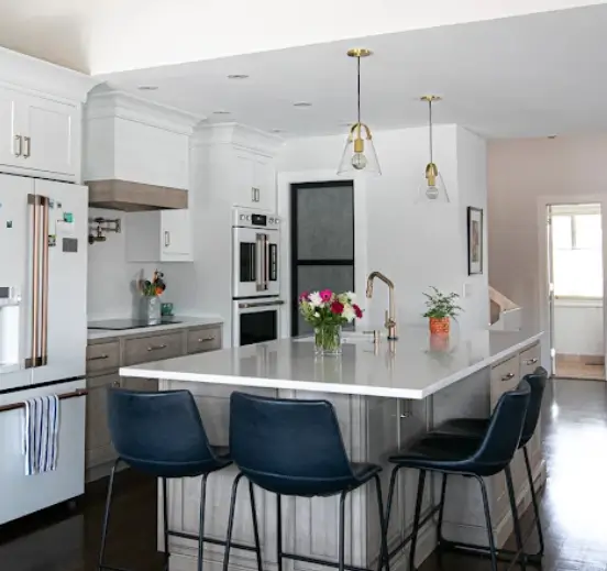 Large kitchen island with integrated seating featuring four dark blue stools, clear glass pendant lights, and a white countertop with flowers.