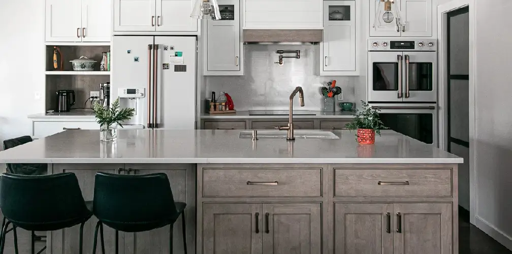 quartzite-vs-quartz-countertops Remodeled Connecticut kitchen island with pale gray engineered stone counter, taupe wood cabinets, gold fixtures, and pendant lighting.