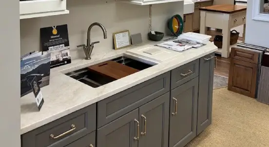 custom-kitchen-cabinetry Showroom display with gray shaker cabinets, brass pulls, a workstation sink with a wood cutting board, and wood door samples in the background.